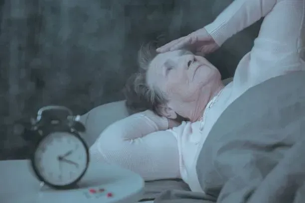 An elderly woman is laying in bed next to an alarm clock.