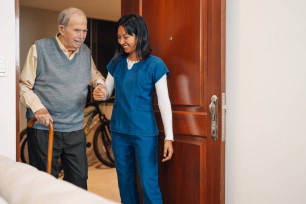 Caregiver assisting an older person with a cane through a doorway.