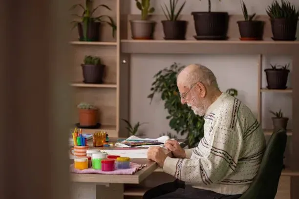 An elderly man is sitting at a desk painting a picture.