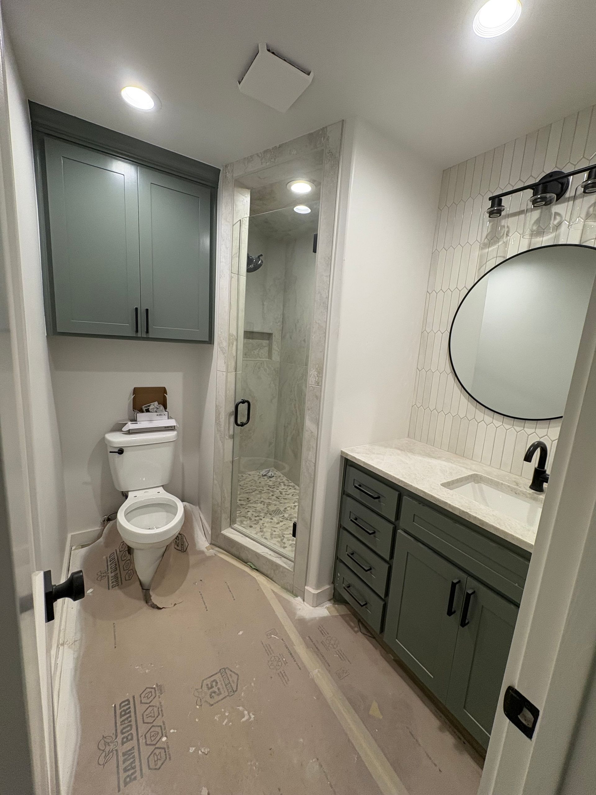 Bathroom with green cabinets, white toilet, and shower. Black fixtures, oval mirror, patterned wall.