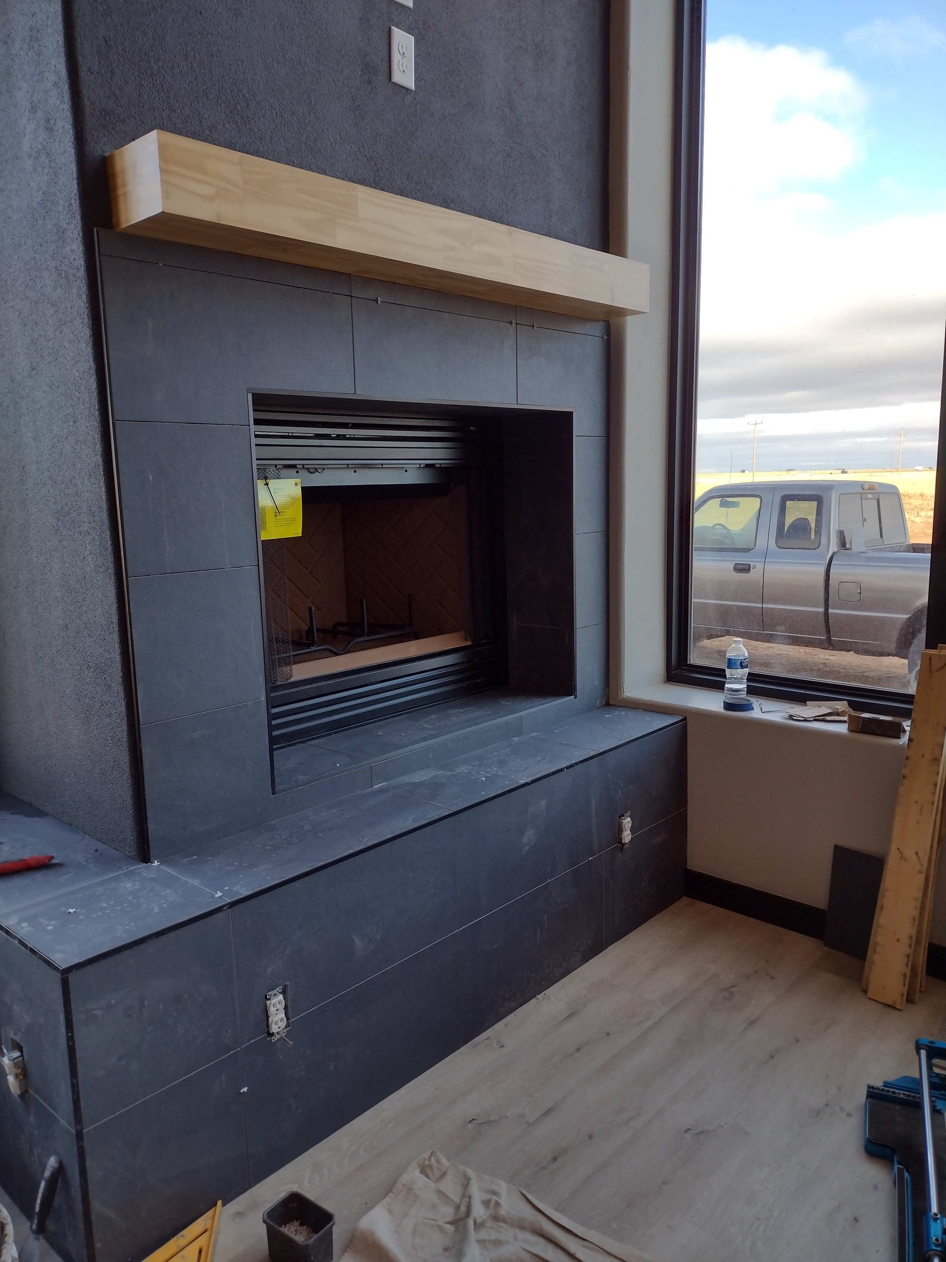 Fireplace under construction, dark gray surround, wood mantel, window with truck visible. Fireplace under construction, dark gray surround, wood mantel, window with truck visible.