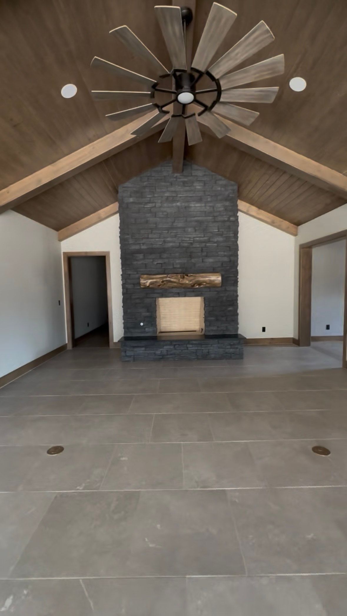 Spacious room with fireplace, wood ceiling, and ceiling fan. Gray tiled floor, neutral walls, and two doorways. Spacious room with fireplace, wood ceiling, and ceiling fan. Gray tiled floor, neutral walls, and two doorways.