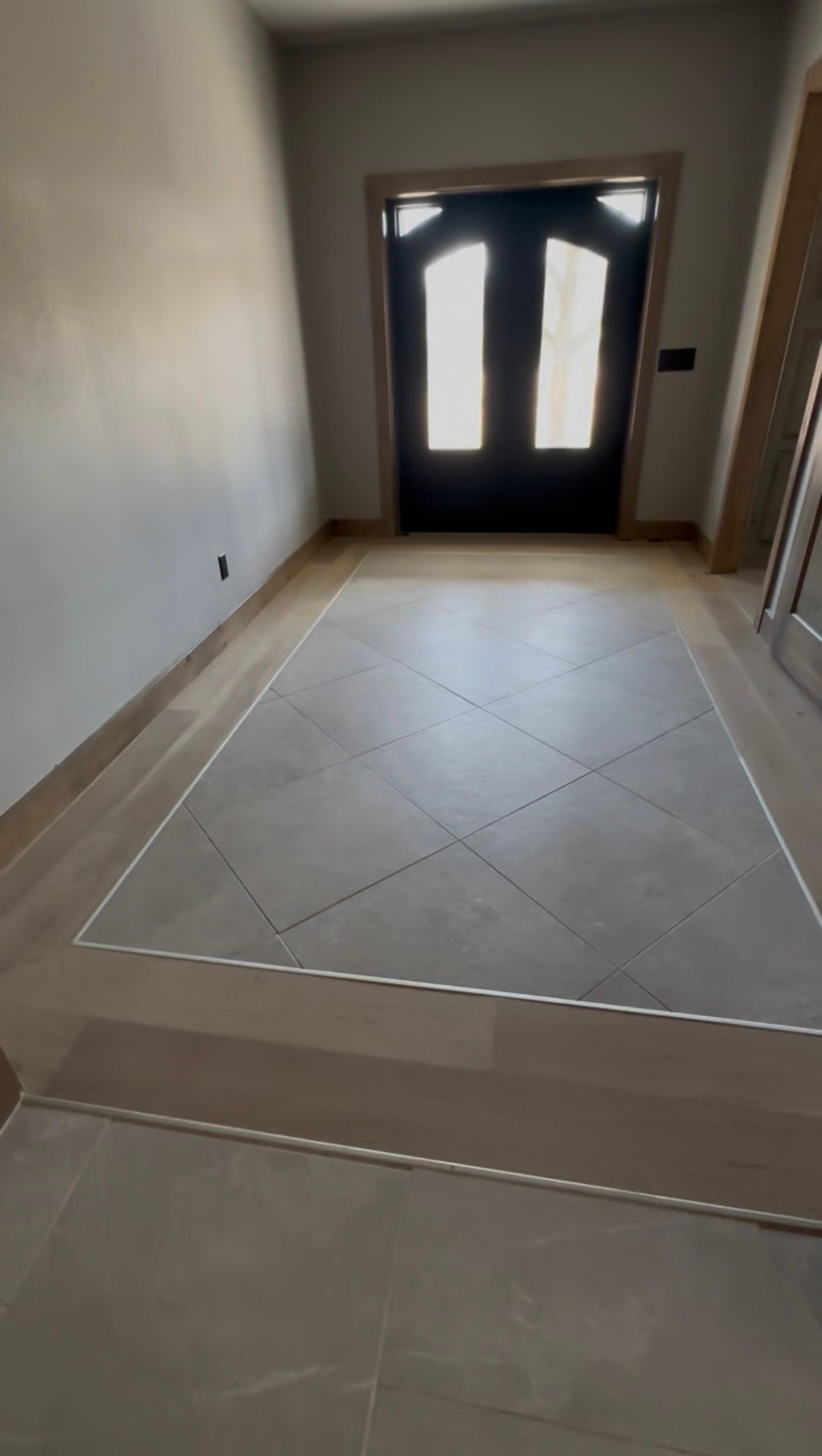 Entryway with light gray tile flooring, a darker gray centered square, and a wood border leading to a black door. Entryway with light gray tile flooring, a darker gray centered square, and a wood border leading to a black door.