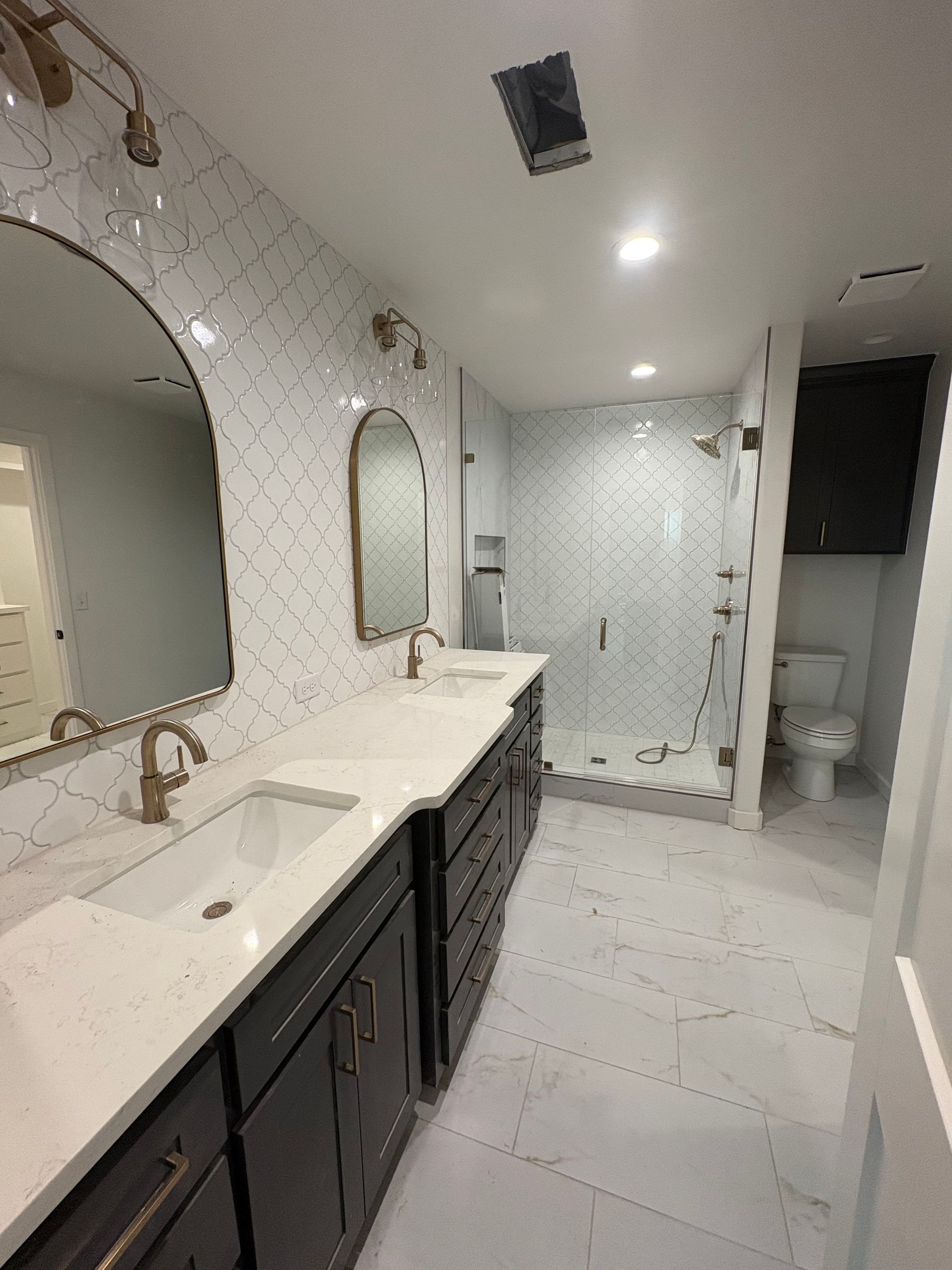 Modern bathroom with white and gold accents, including dual sinks, arched mirrors, and a glass shower.