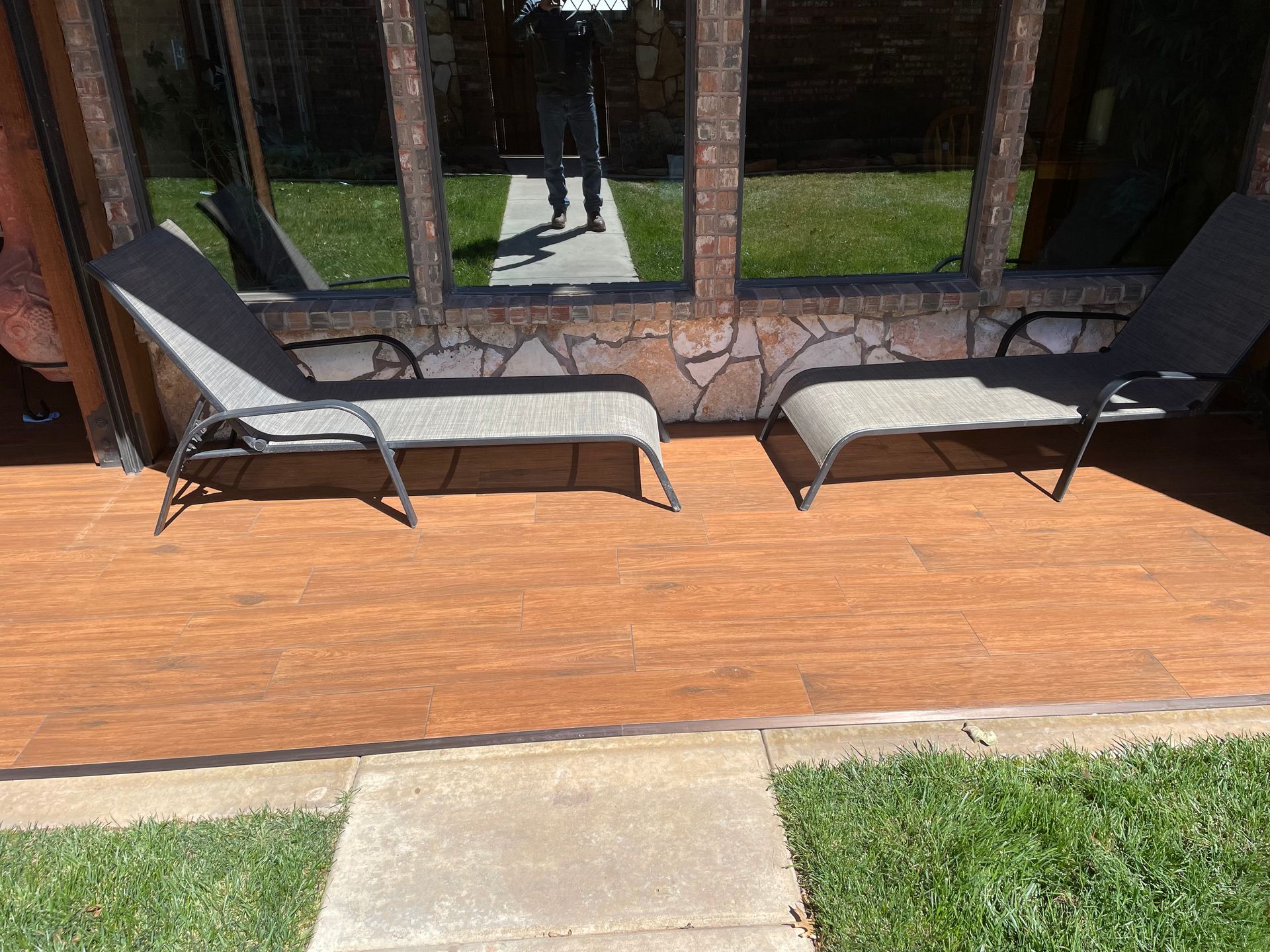 Two gray chaise lounges on a wood-look deck outside a building with windows. A person is reflected in the windows.