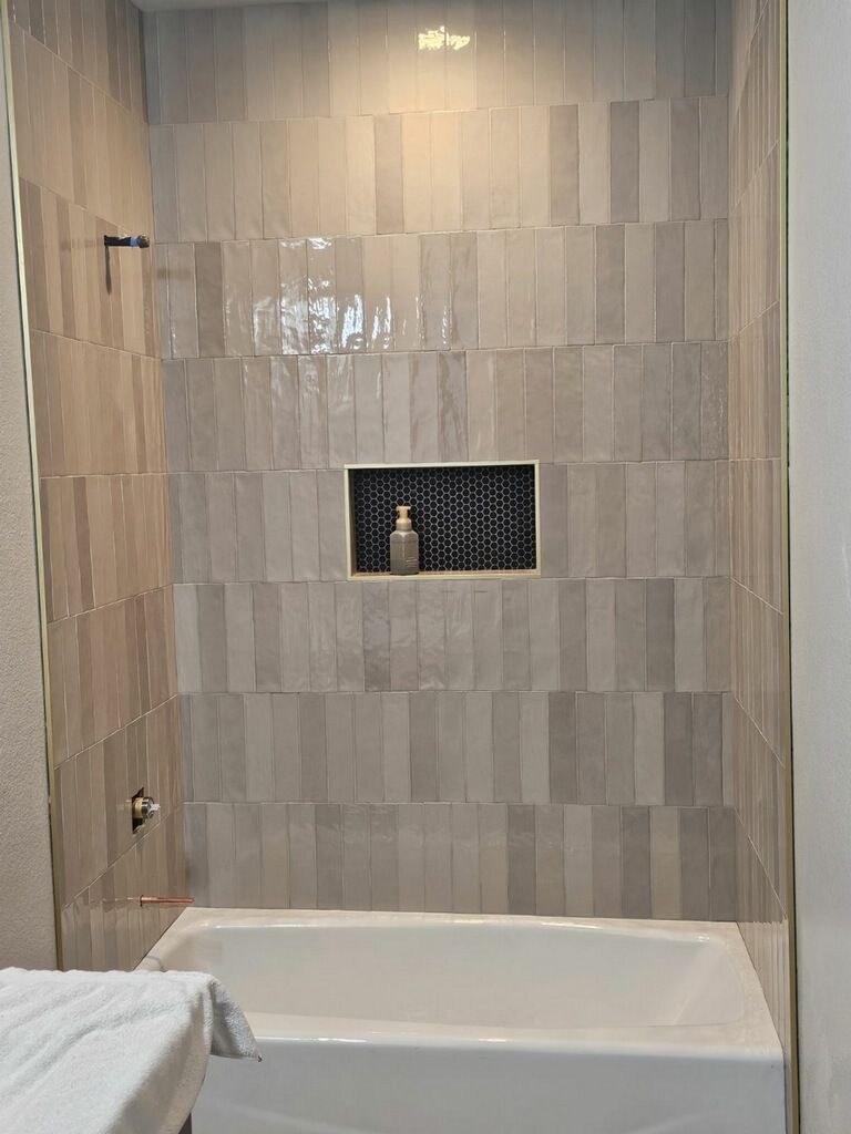 Bathroom with a white bathtub and vertically stacked grey tiles; a recessed shelf holds a bottle.