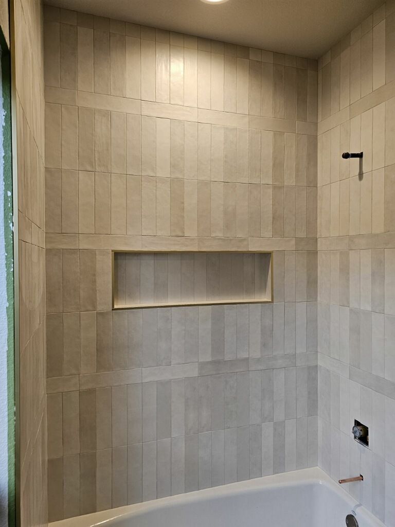 Tiled shower interior with a built-in niche. Vertical, light gray tiles cover the walls. A faucet and showerhead are visible.