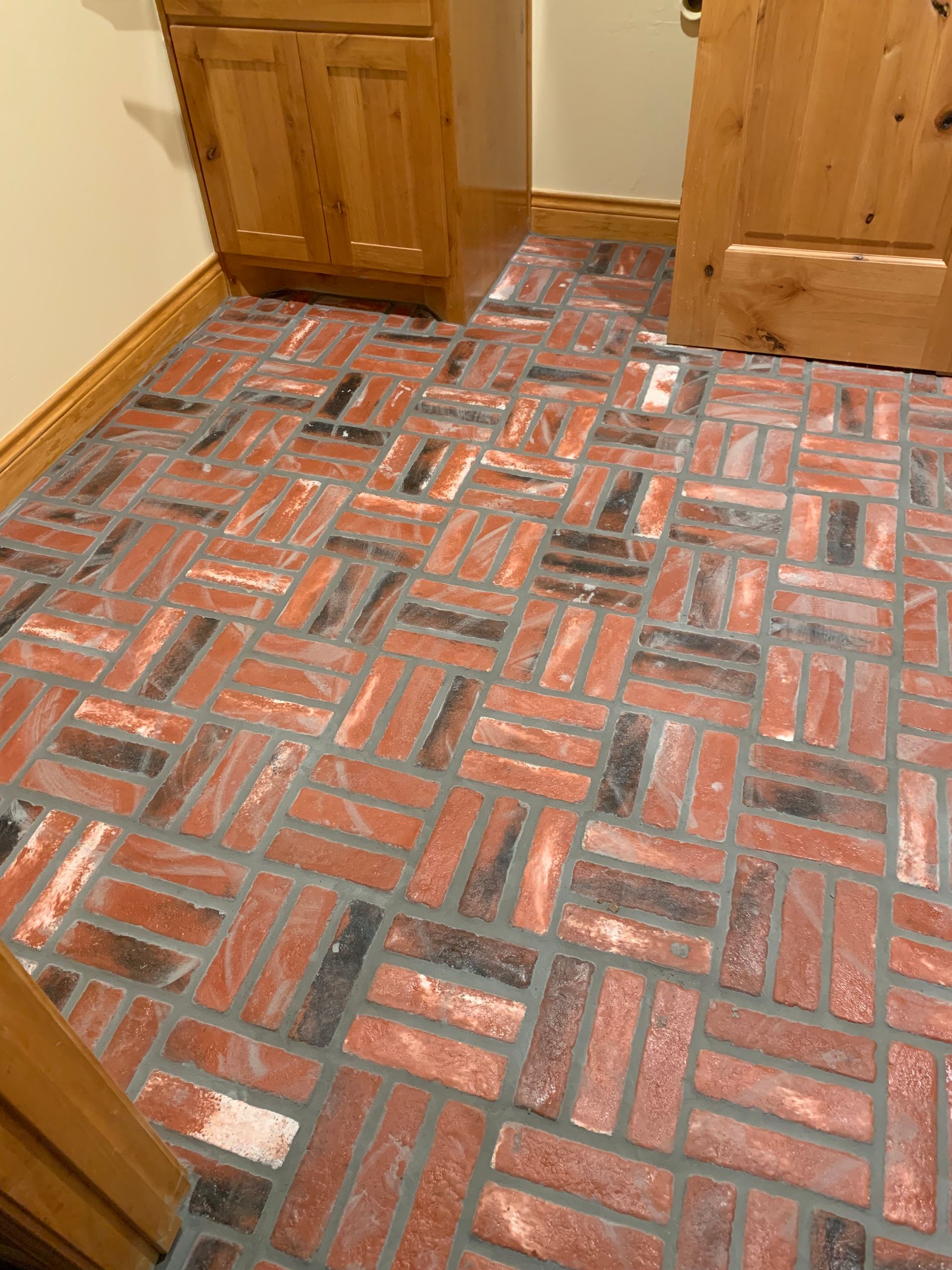 Brick flooring in herringbone pattern, red and black bricks with gray grout, inside a room with wooden cabinets and a door. Brick flooring in herringbone pattern, red and black bricks with gray grout, inside a room with wooden cabinets and a door.