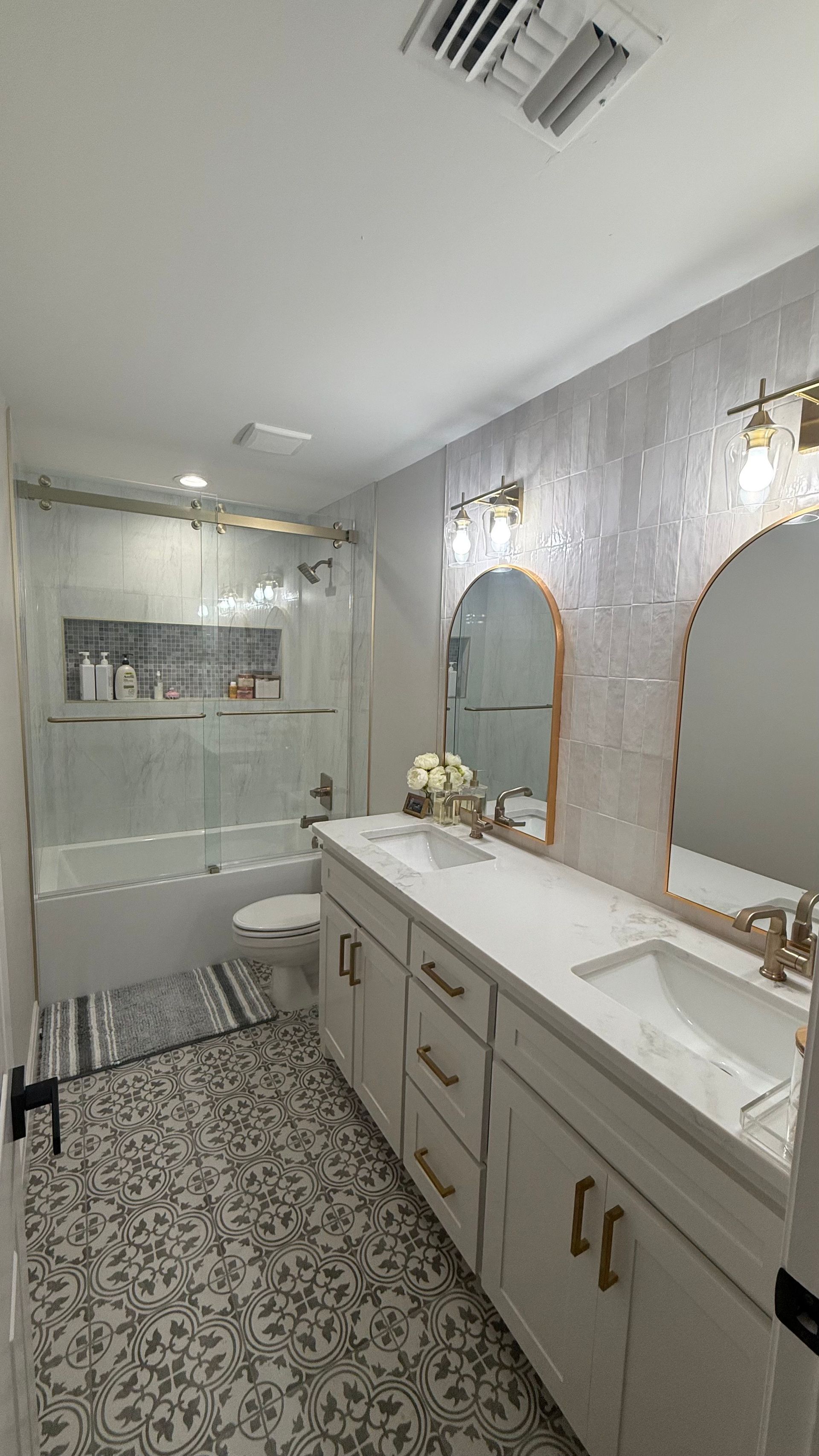 Bathroom with dual sinks, shower, and patterned floor. White cabinets, gold fixtures, and textured wall.