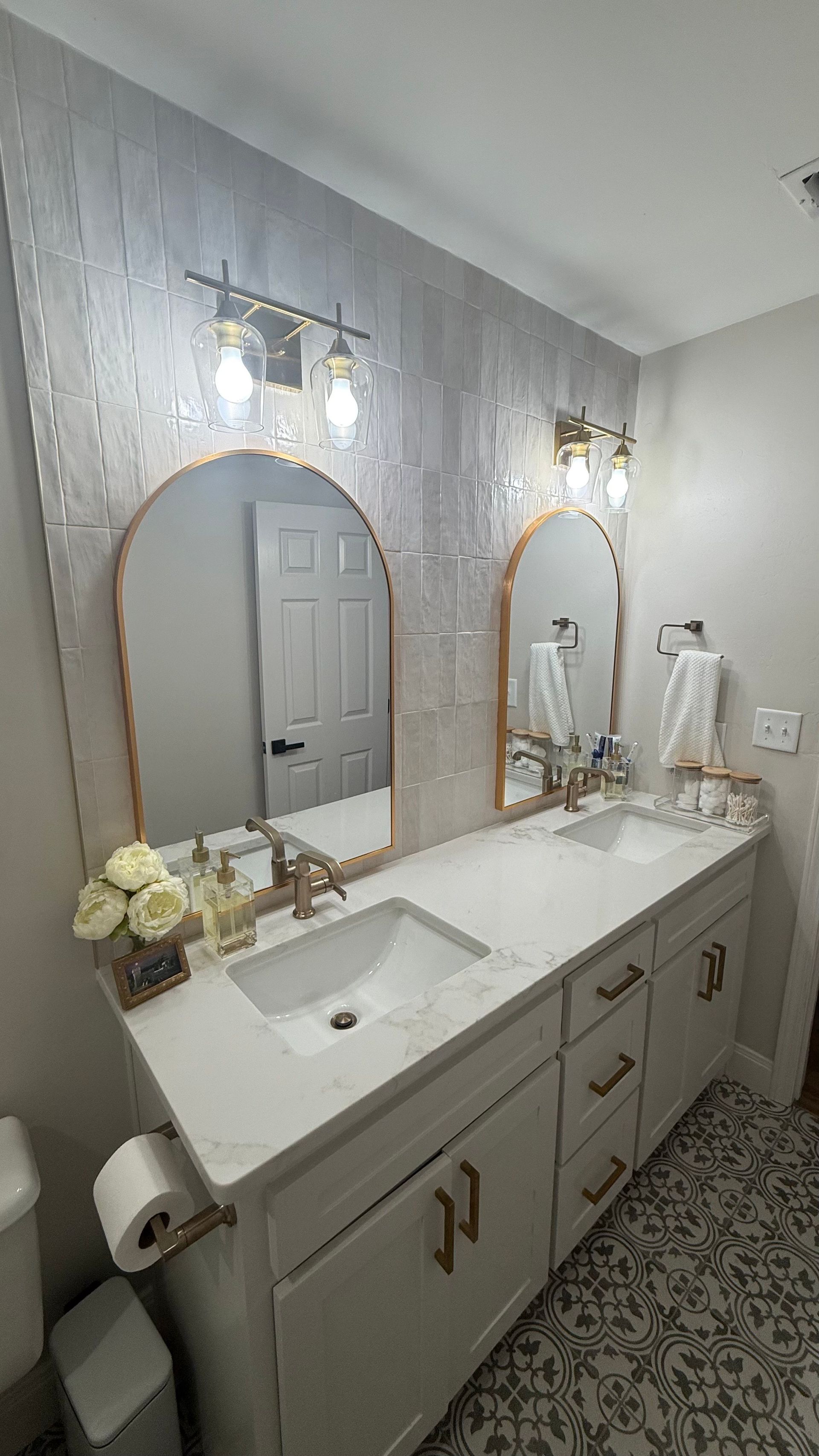 Double vanity bathroom with arched mirrors, white marble countertops, gold hardware, and patterned floor.