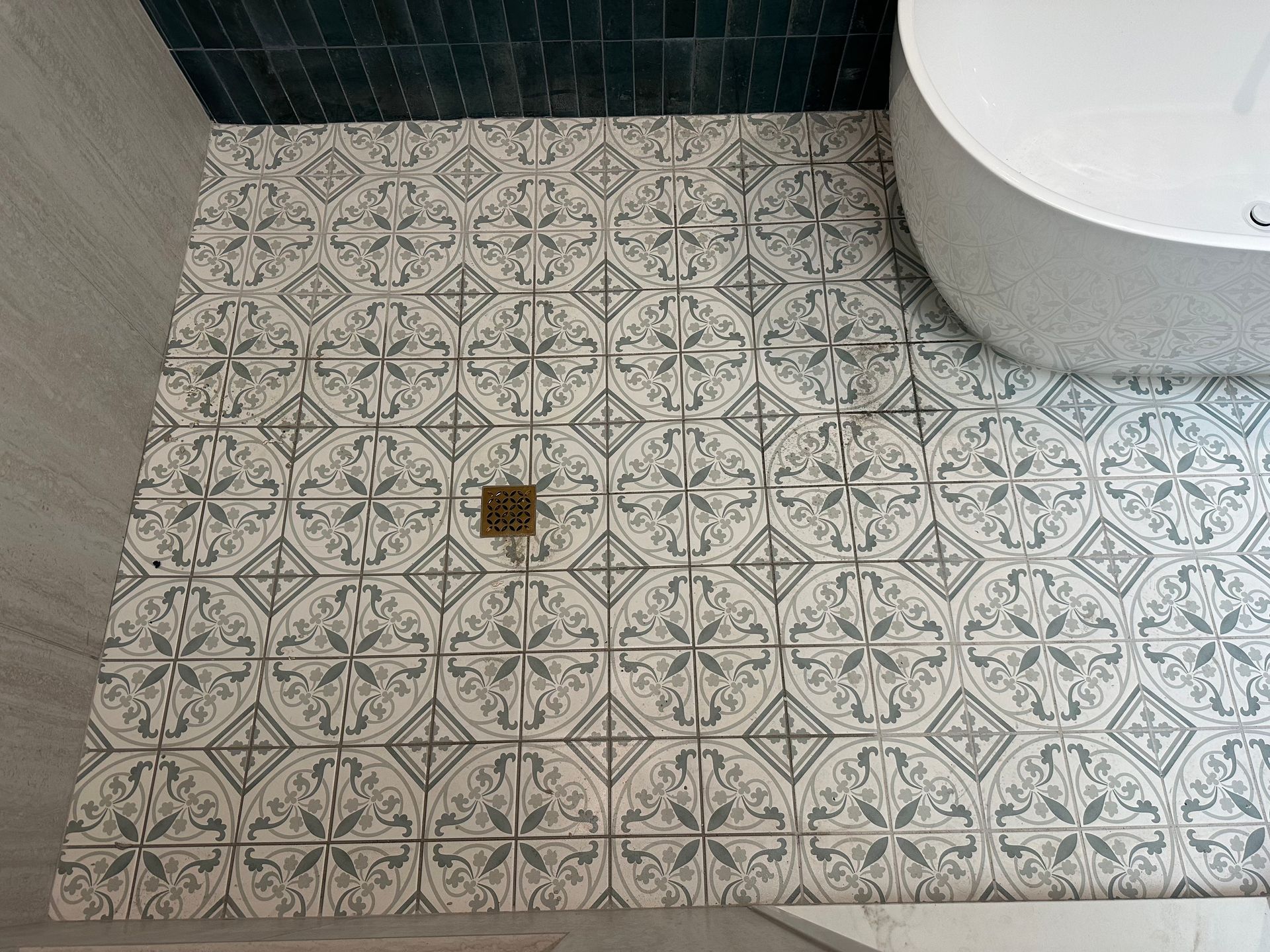 Bathroom floor with ornate gray and white tiles, gold drain, and white tub. Dark teal tiles on the wall. Bathroom floor with ornate gray and white tiles, gold drain, and white tub. Dark teal tiles on the wall.