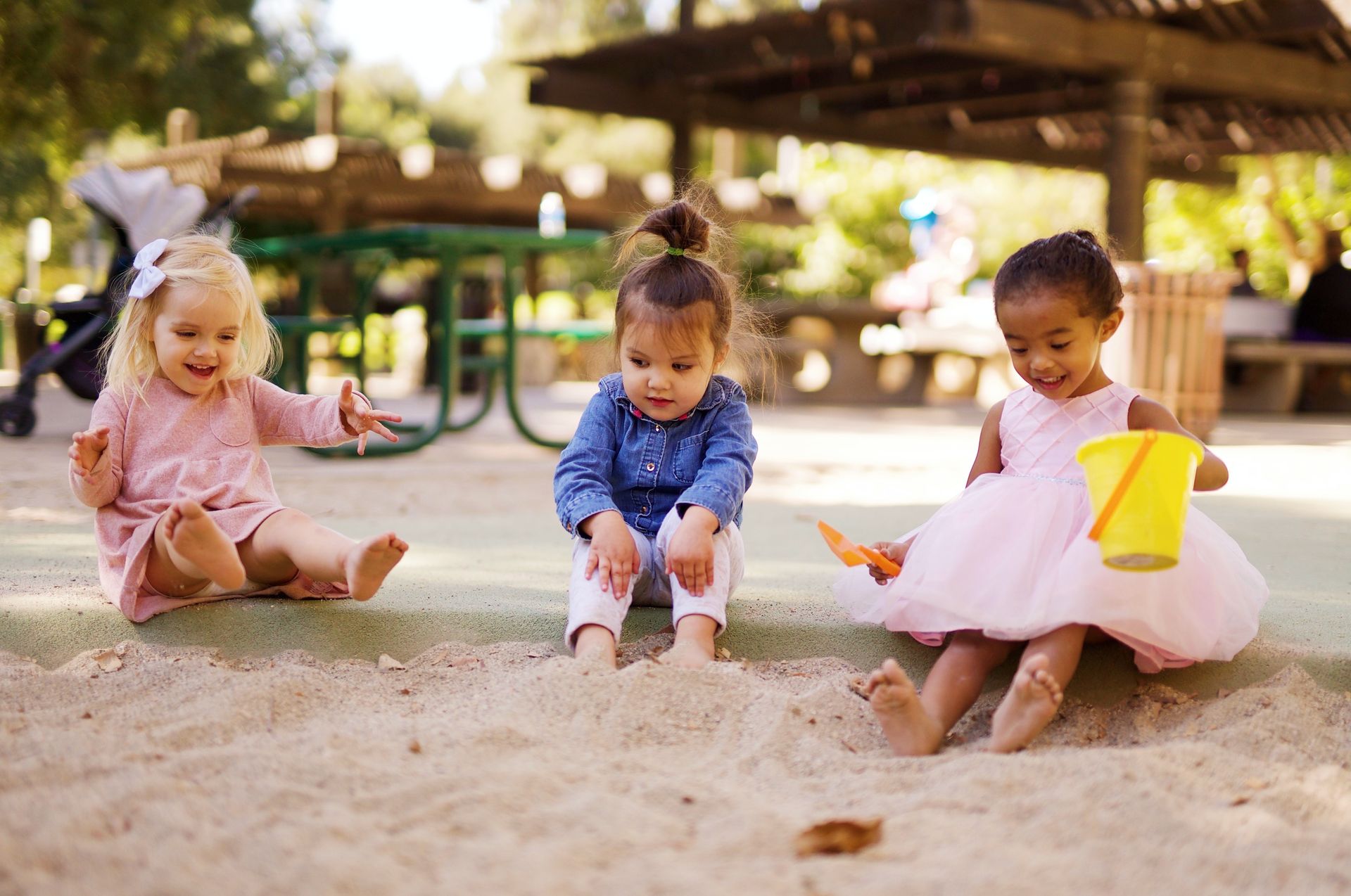 Three children playing in a sandbox outdoors, sitting with colorful buckets and shovels.