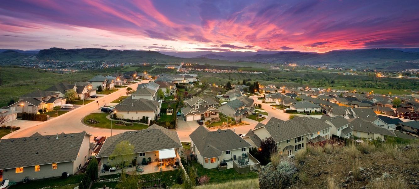 Sunset over a suburban neighborhood with winding streets, houses, and distant mountains under a pink sky