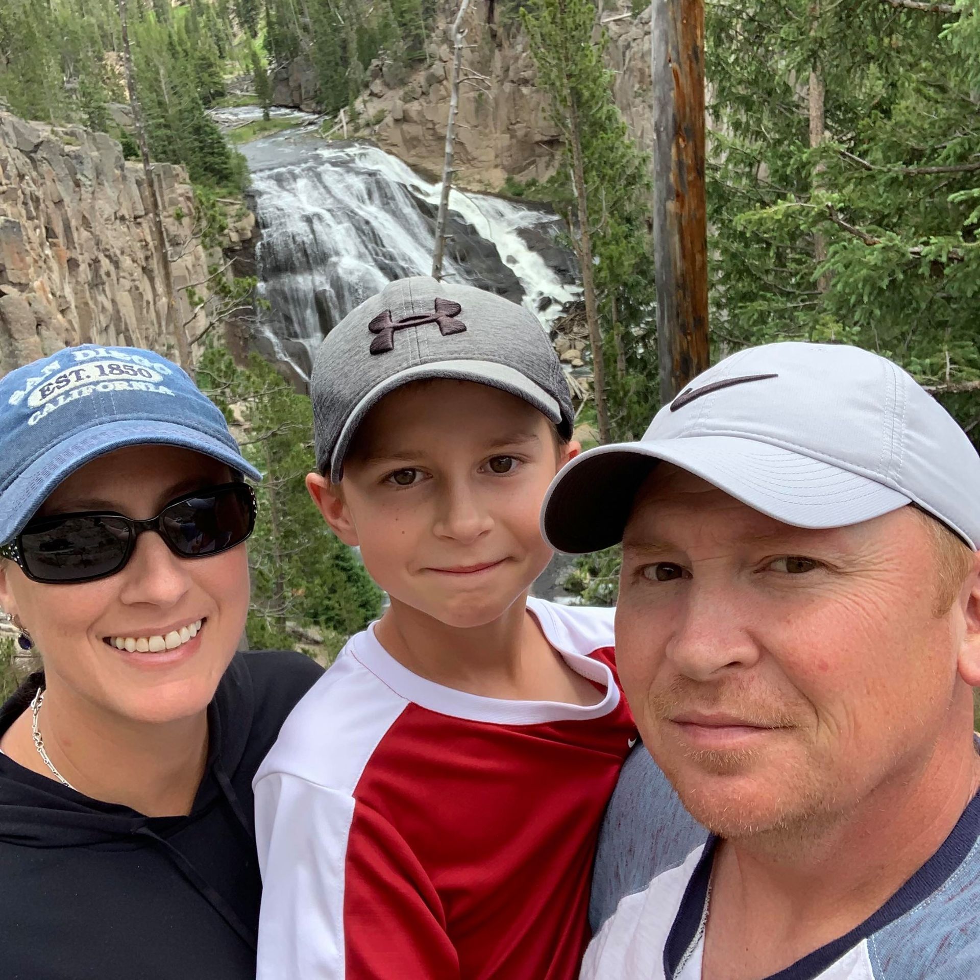 Family selfie in front of waterfall; woman in sunglasses, boy in red, man in hat.