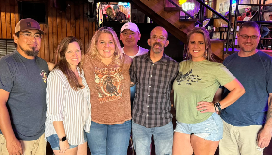 Group of seven people smiling for a photo inside a dimly lit bar, with a wooden interior and some neon signs visible.