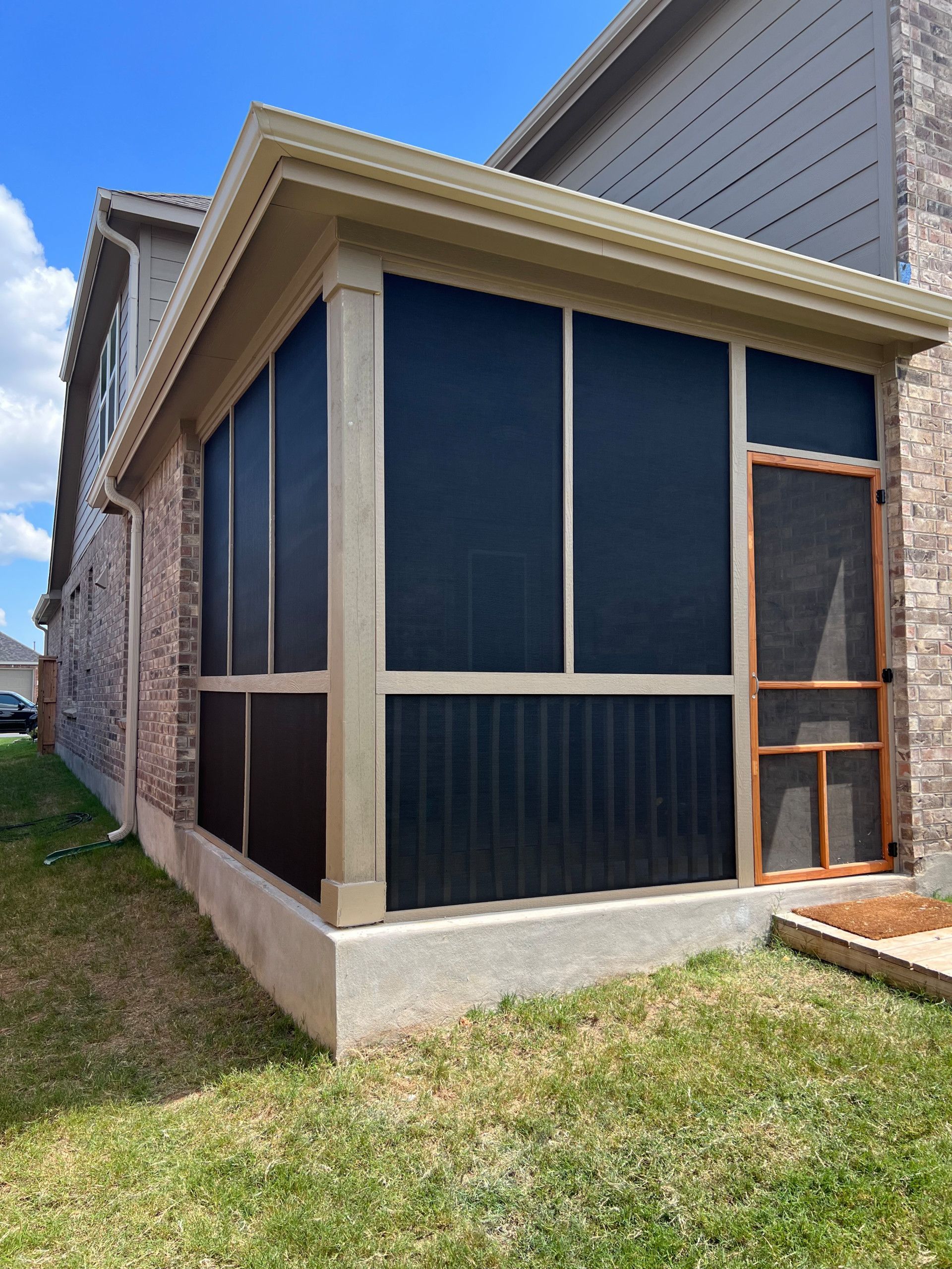A screened-in porch attached to a brick house. The screens are dark, with tan trim and a concrete base.