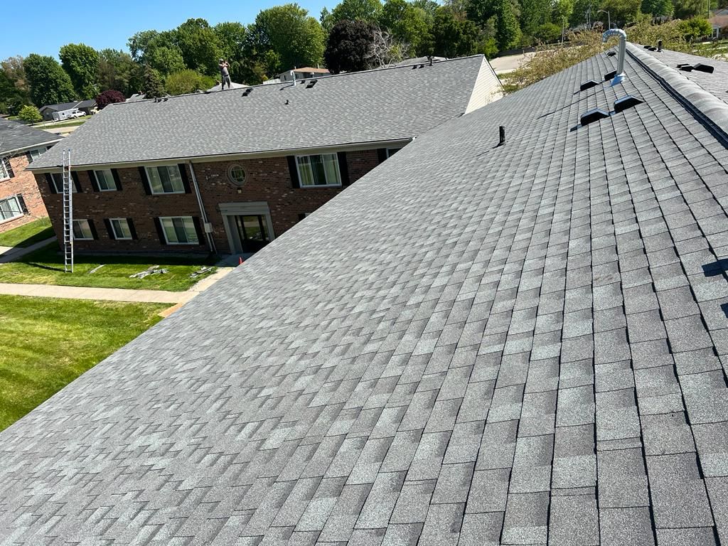 View across a gray shingled roof to a brick apartment building and trees on a sunny day