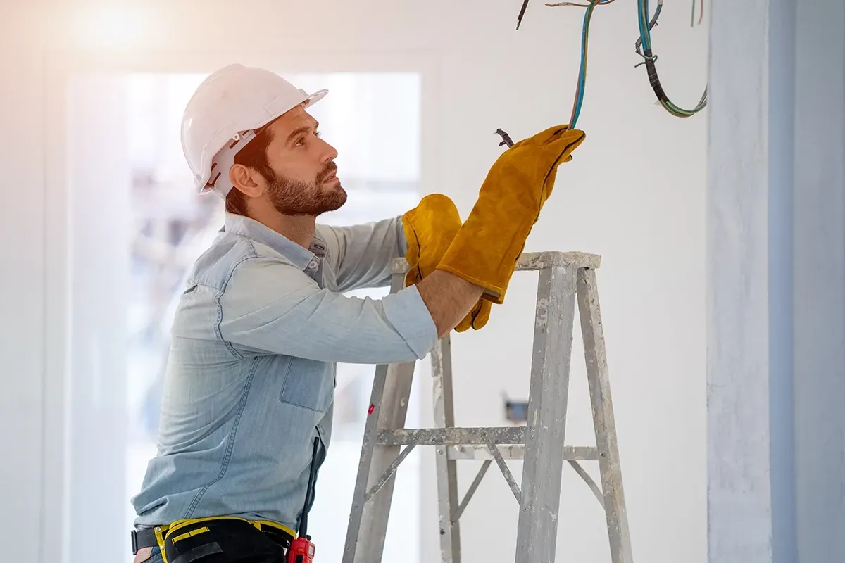 An electrician in a hard hat and protective gloves stands on a ladder, inspecting electrical wires in an indoor space.