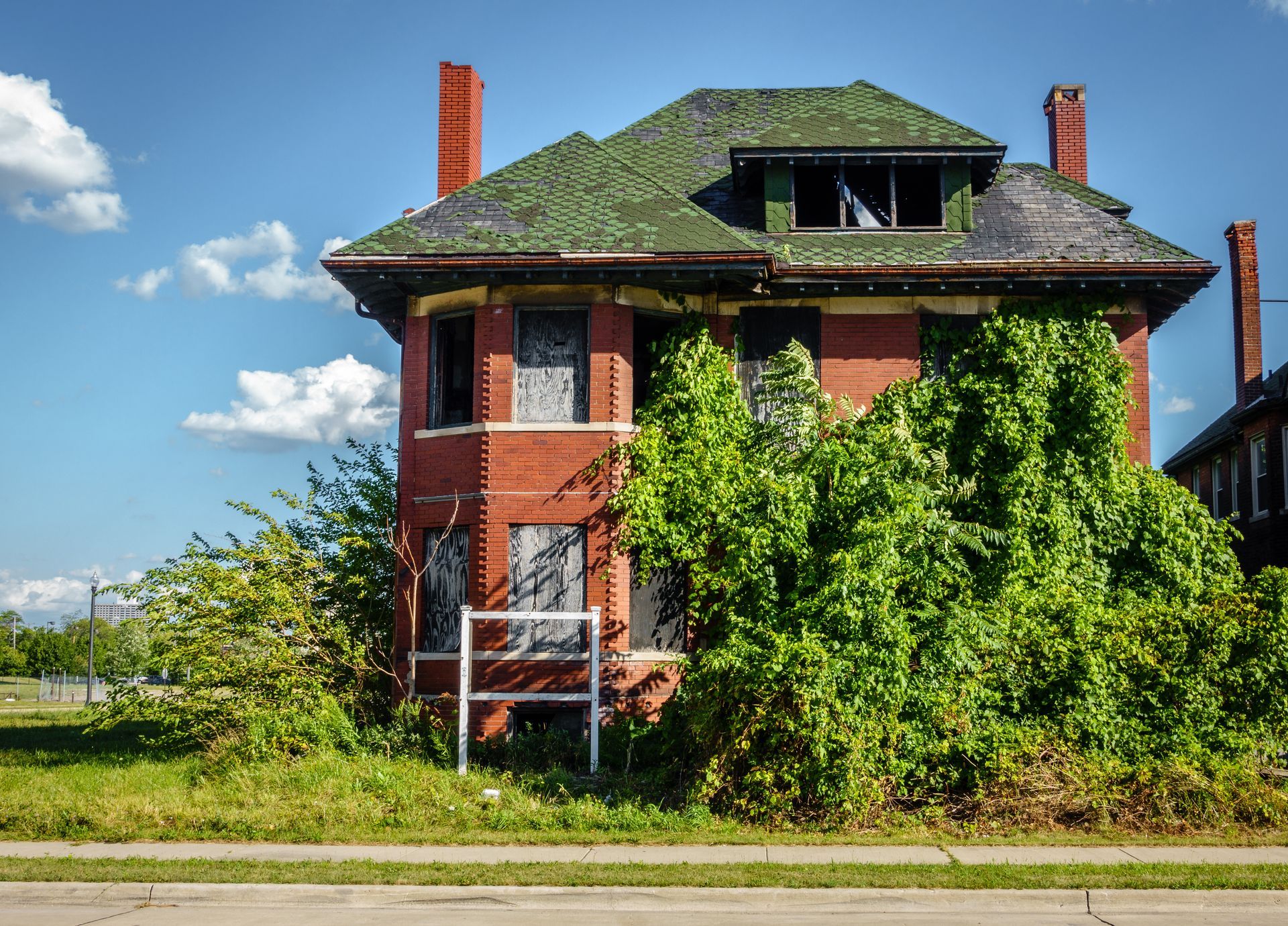 An abandoned brick house with a green roof is surrounded by trees and grass.