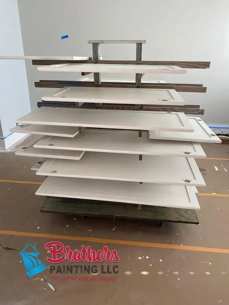 Stacked painted cabinet doors and panels drying in a workshop, with Bruders Painting LLC logo at bottom.