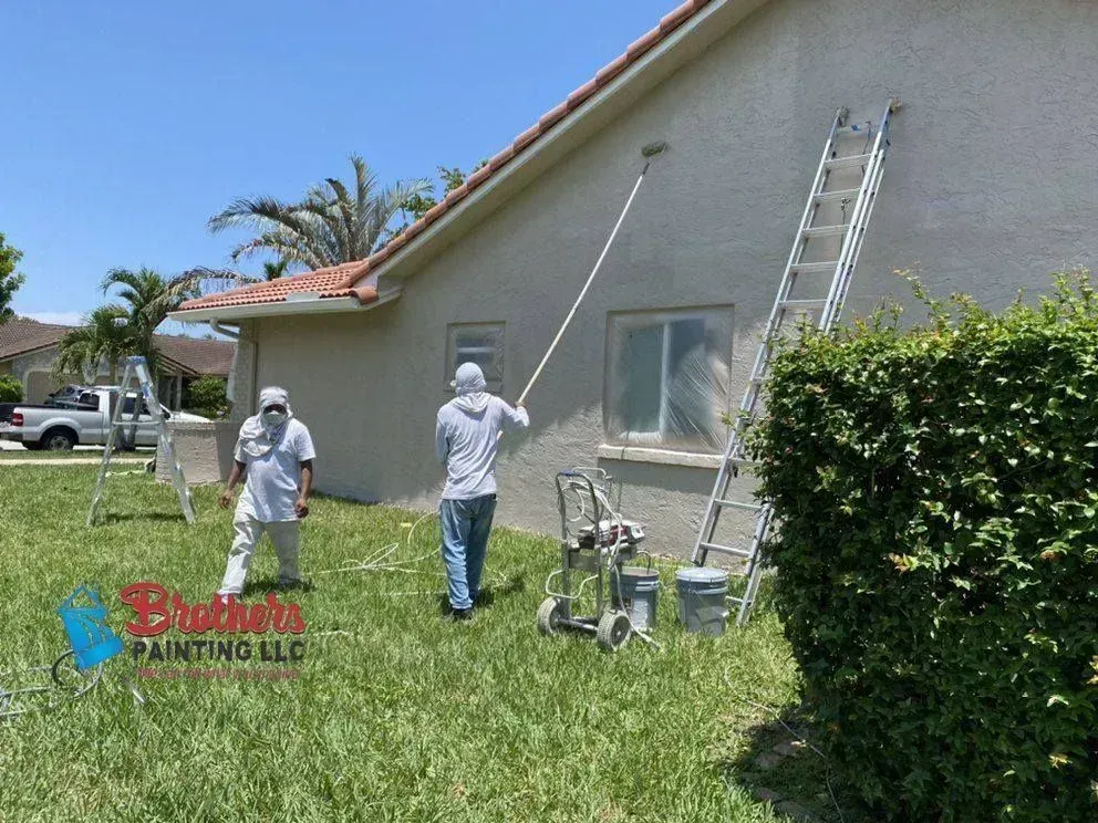 Two workers painting a house exterior with spray equipment and ladders in a sunny yard
