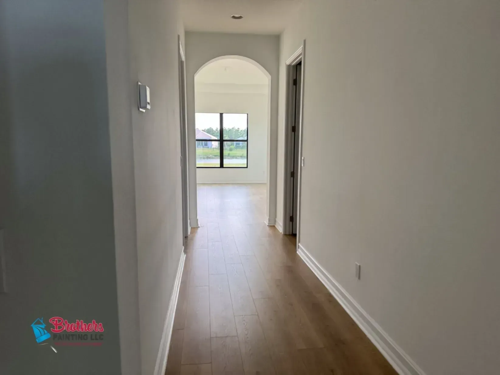 Long white hallway with arched doorway, light wood floors, and a view of a bright room and window at the end