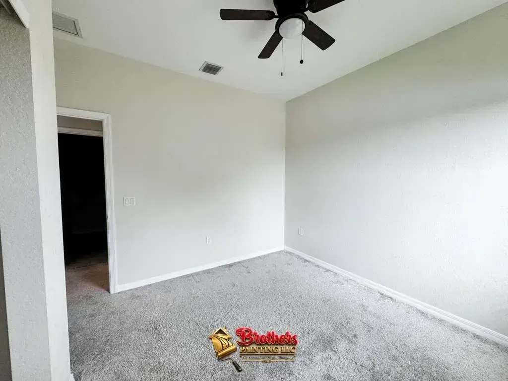Empty beige bedroom with gray carpet, white walls, ceiling fan, and open doorway