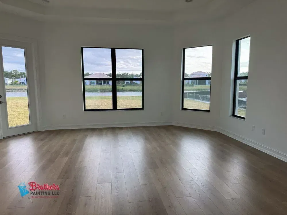 Empty modern living room with light wood floors, white walls, and large windows overlooking a yard