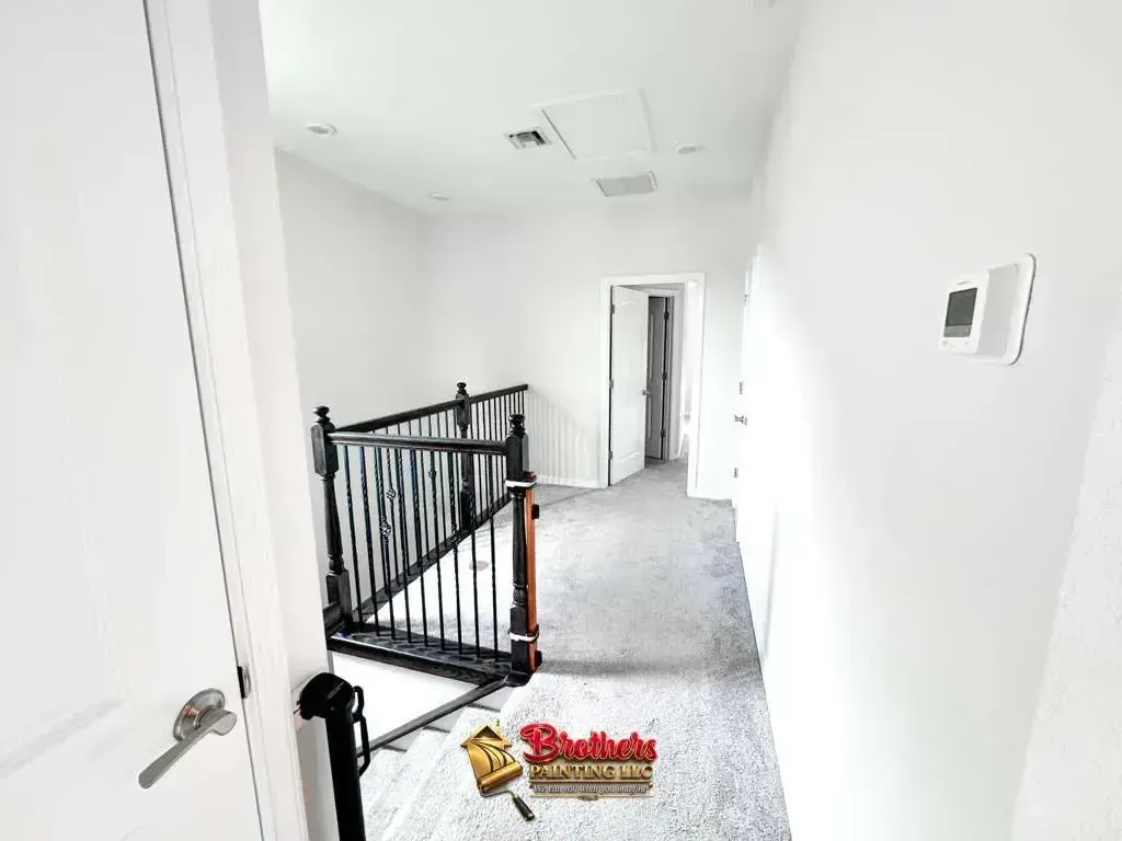 Bright white hallway with black stair railing, gray carpet, and wall thermostat