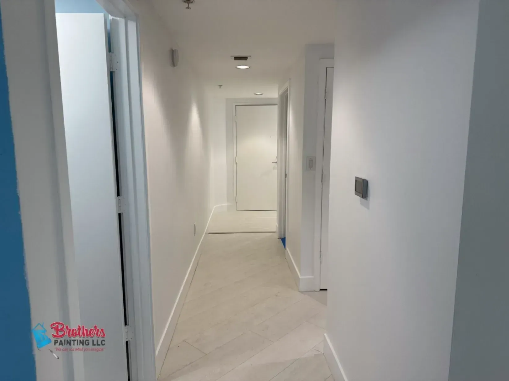 Bright white hallway with doors and recessed ceiling lights, leading to a closed door at the end.