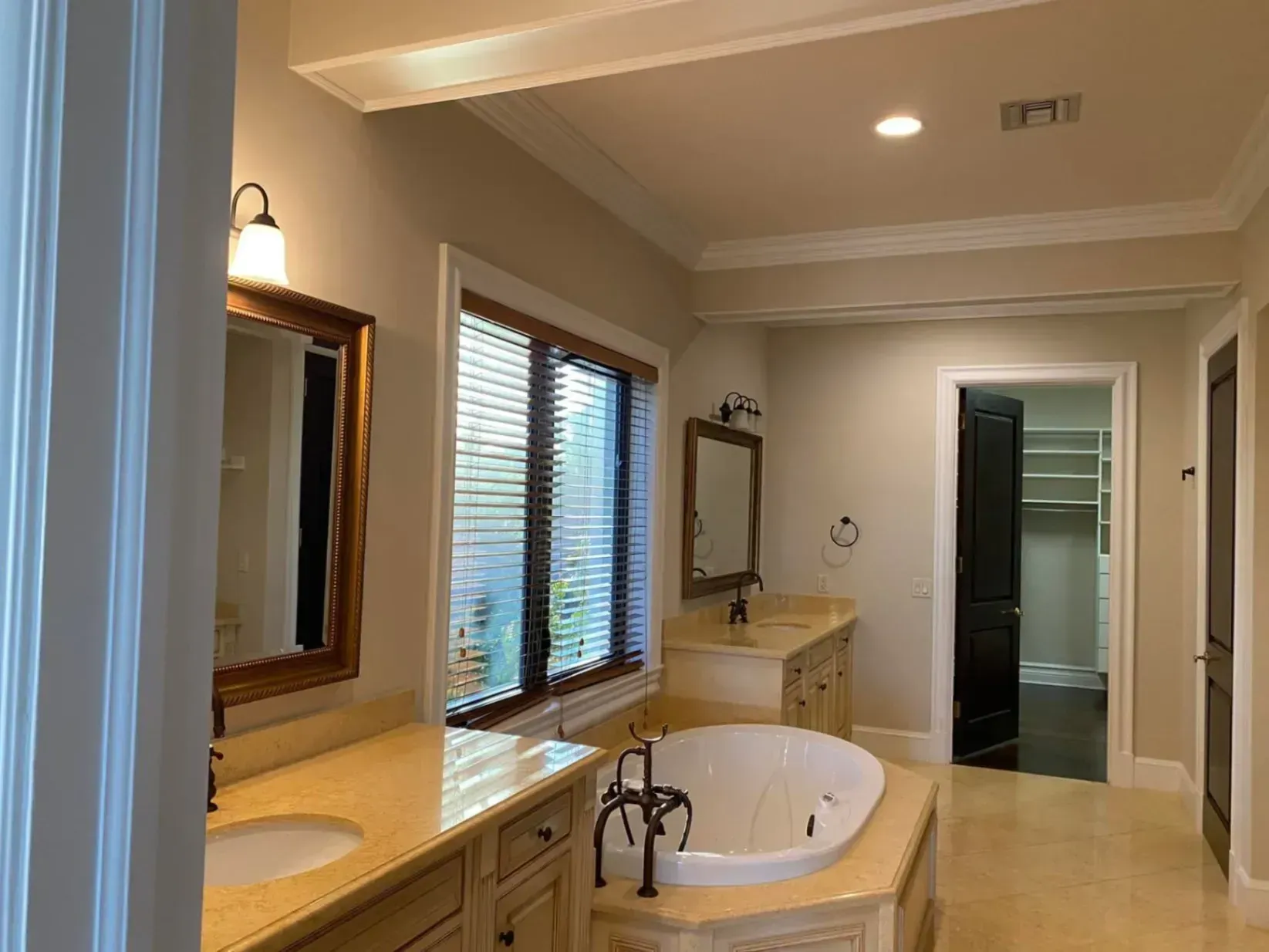 Elegant bathroom with a soaking tub, double vanity, mirrors, and window blinds under warm lighting