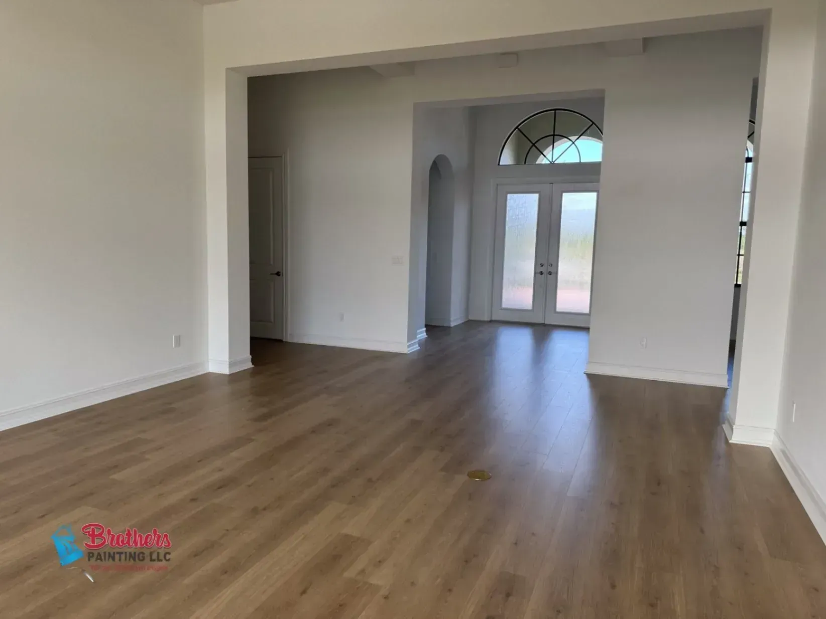 Empty modern living room with wood floors and double front doors under an arched window.