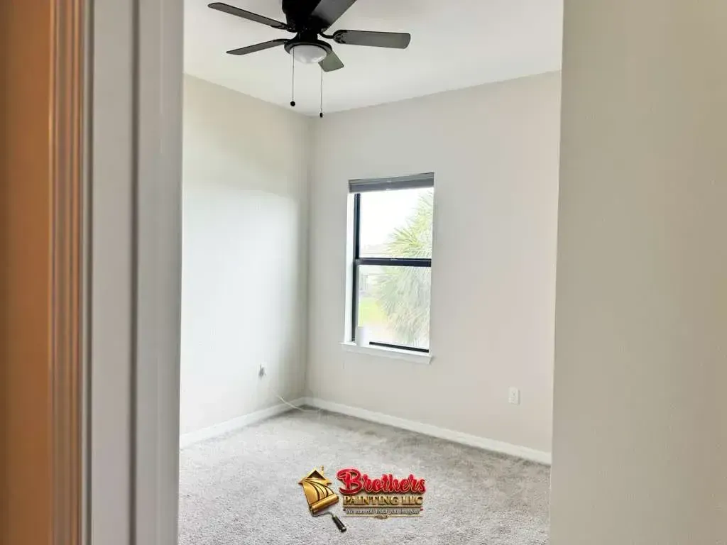 Empty beige bedroom with white carpet, ceiling fan, and a single window.