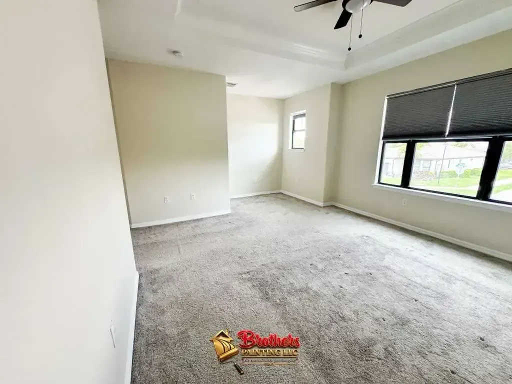 Empty beige bedroom with carpet, ceiling fan, and windows with black blinds.