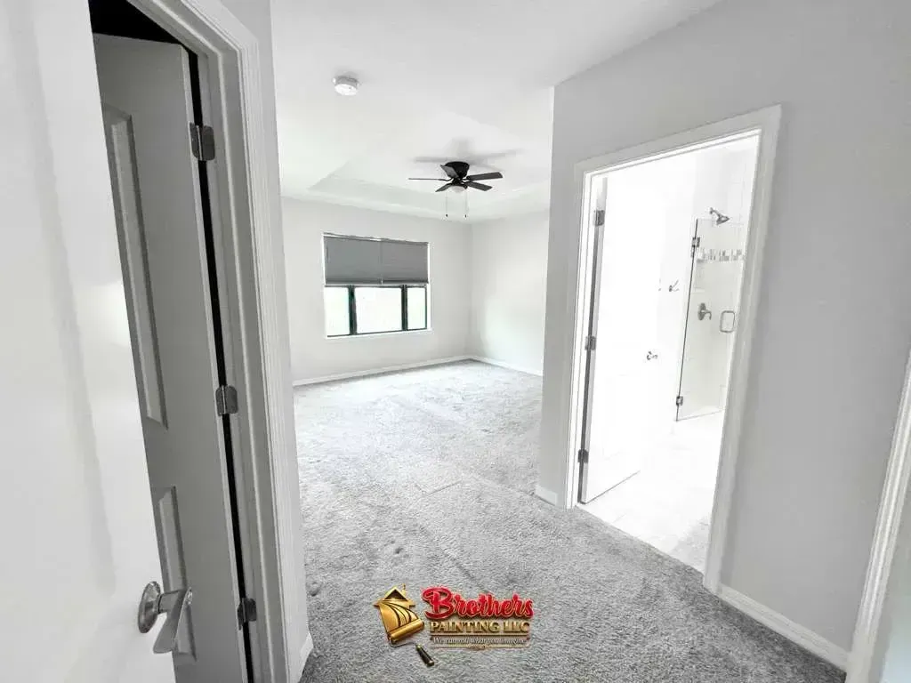 Bright empty bedroom with gray carpet, white walls, ceiling fan, and open bathroom doorway