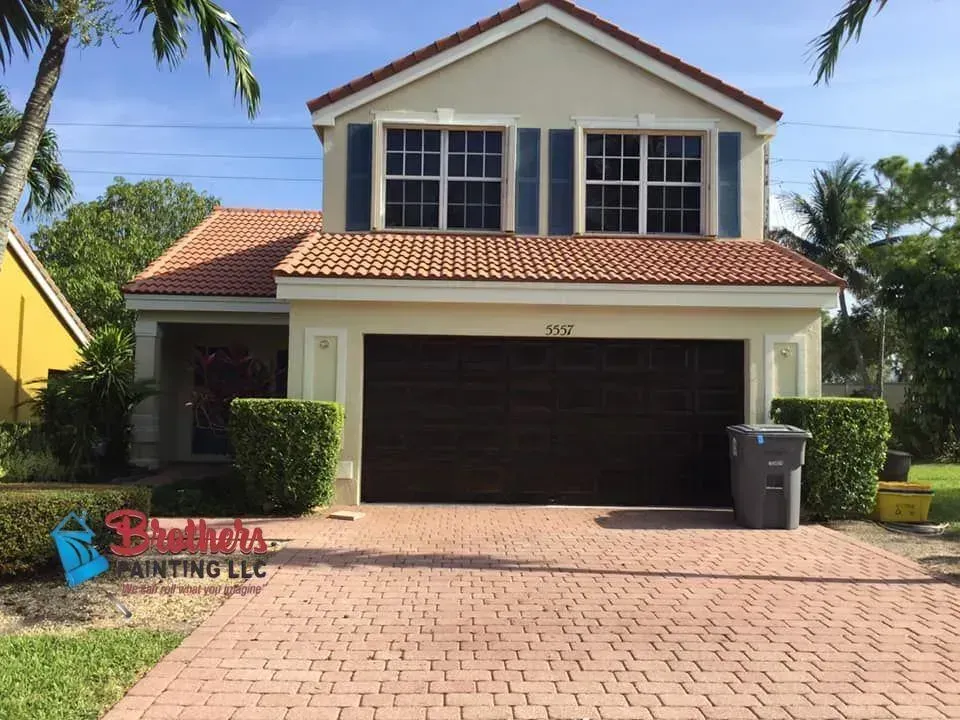 Two-story beige house with red tile roof, dark garage door, and brick driveway under palm trees