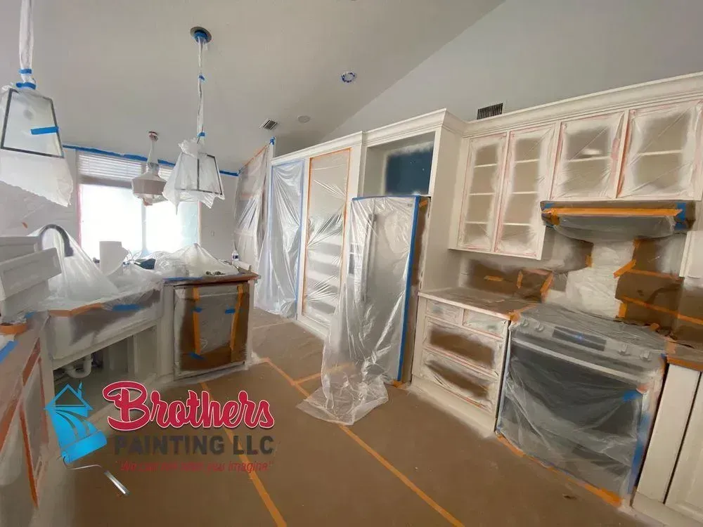 Kitchen under renovation with white cabinets, plastic-covered appliances, and painting supplies on the floor