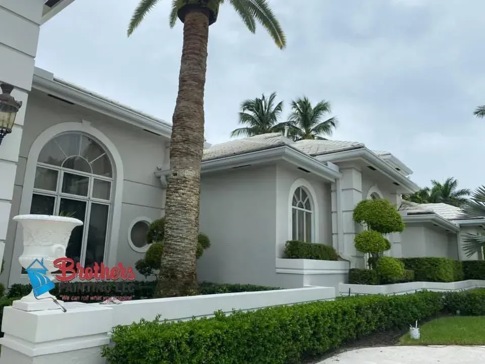 White Mediterranean-style house with arched windows, palm trees, and manicured hedges under a cloudy sky