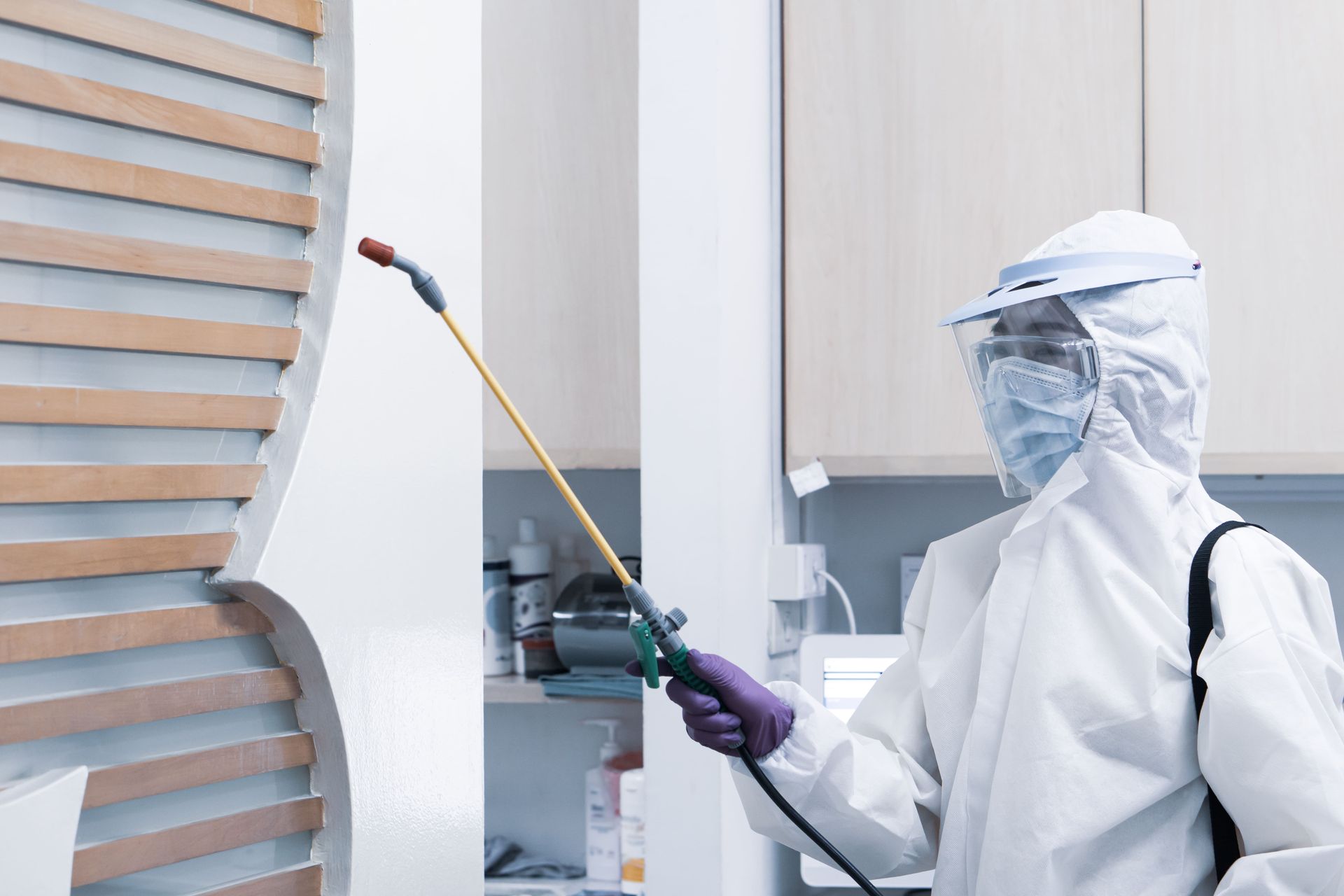 A person in full white protective gear and a face shield sprays disinfectant on a wooden paneled wall in an indoor space.