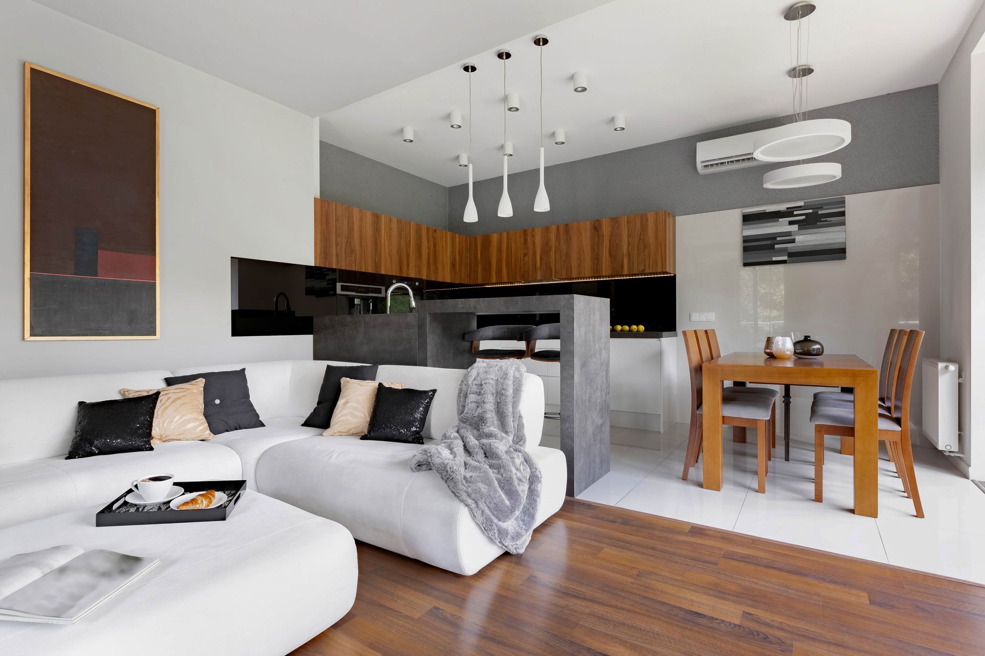 A modern open-concept living area with a white sectional sofa, wooden dining table, and kitchen with wood-toned cabinets.
