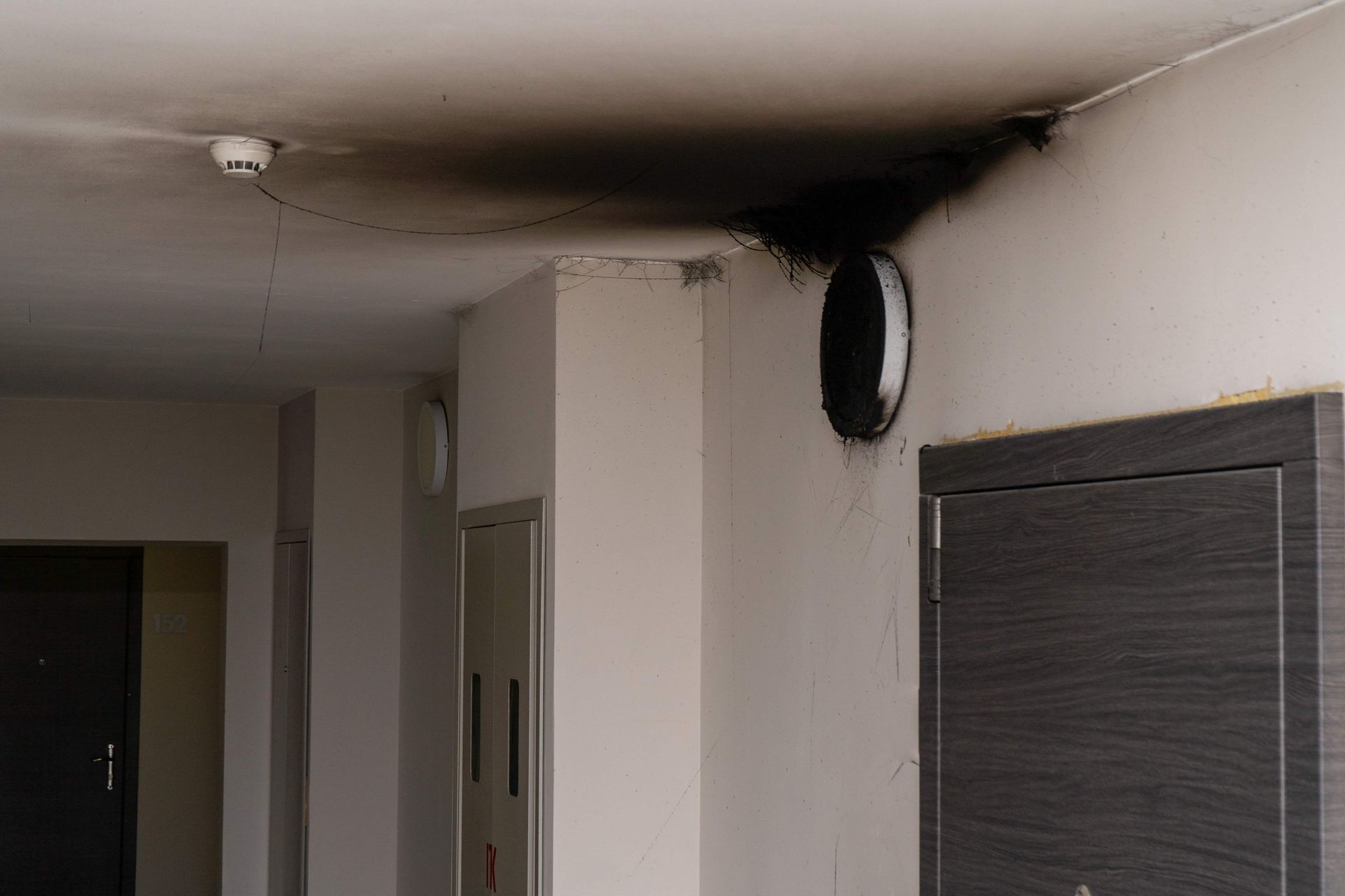 A dark soot stain spreads across a white ceiling and wall, surrounding a circular black vent in an indoor hallway.