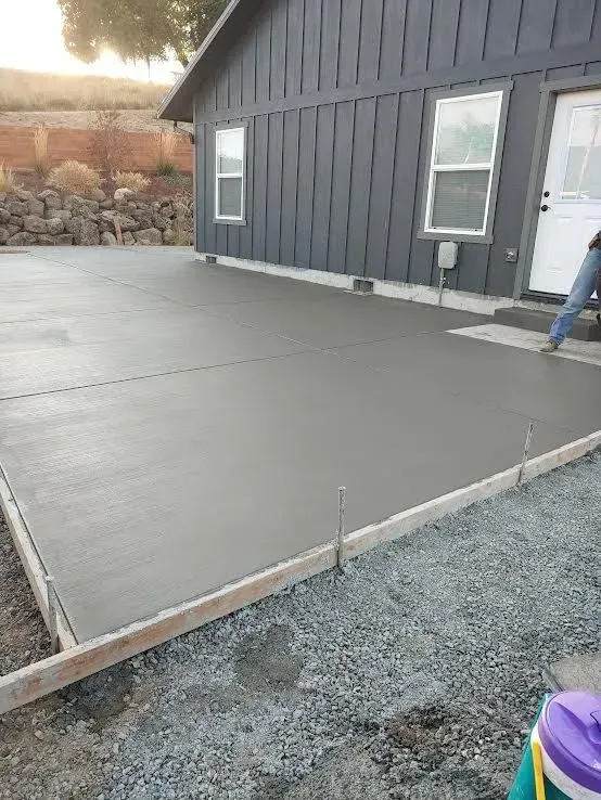 Fresh gray concrete patio beside a dark house, with gravel edge and a worker partially visible on the right.