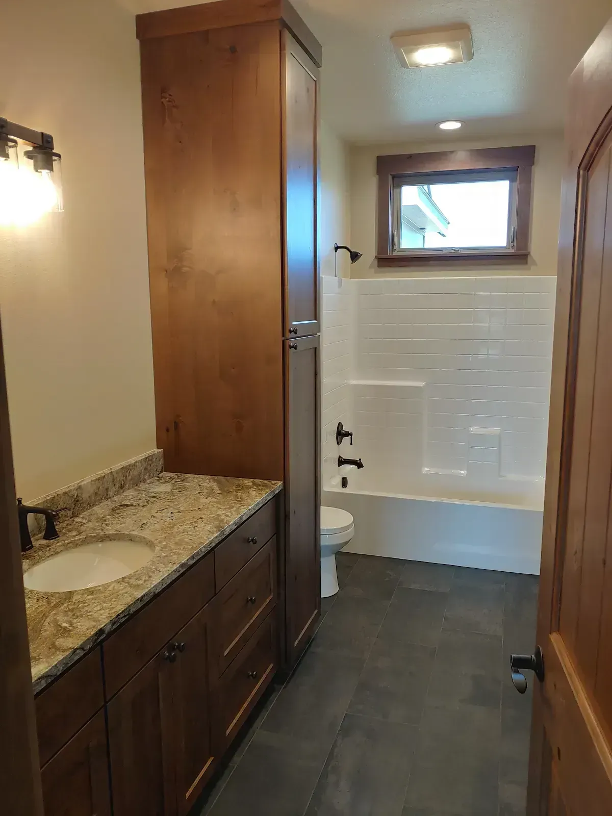 Narrow bathroom with dark wood vanity, granite countertop, and white tub/shower with window.