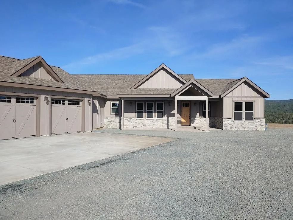 Single-story beige house with three garage doors, gravel driveway, and covered front porch under a blue sky