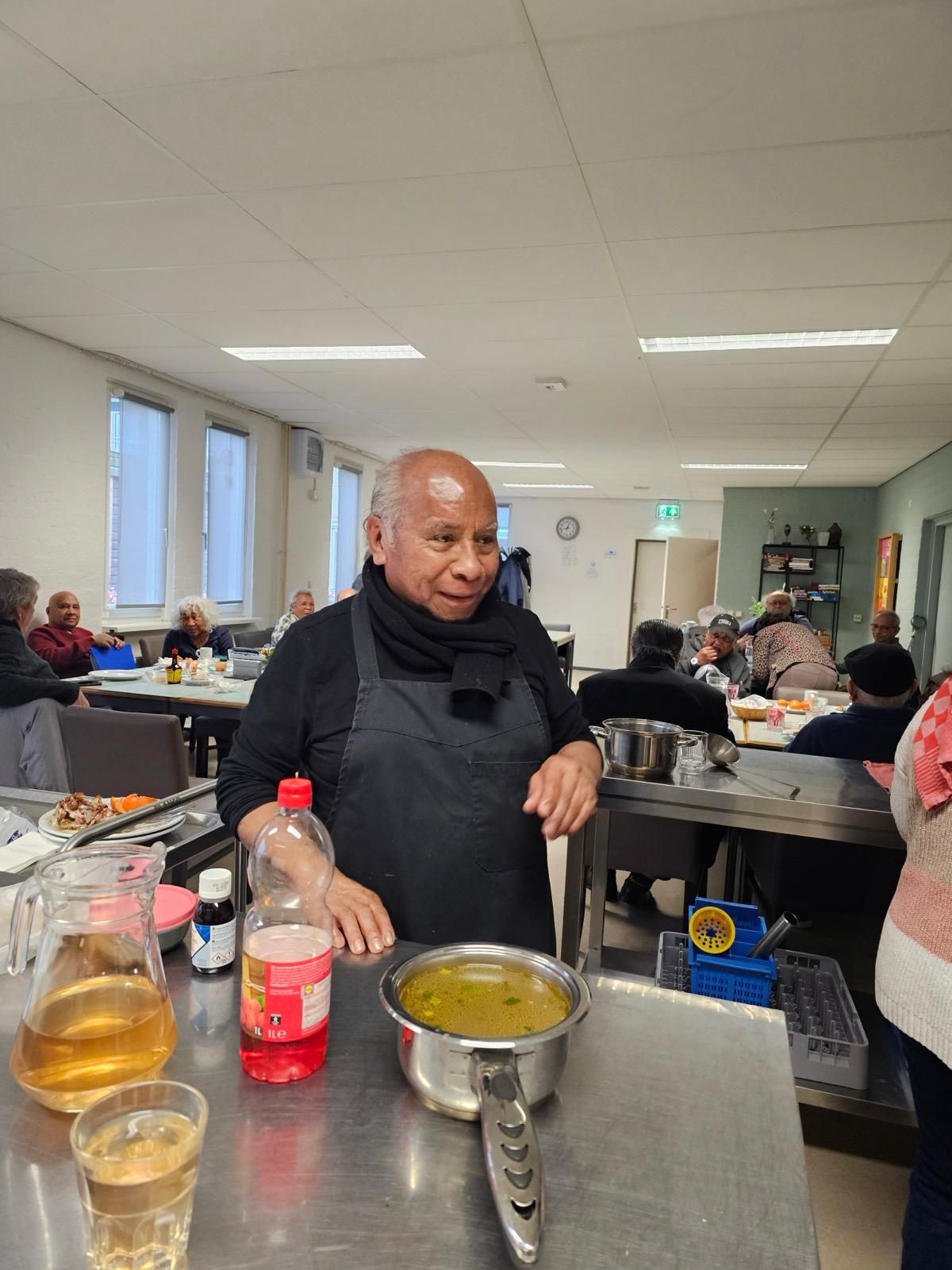 Bij de toonbank staat een man met een schort aan met een kom soep en een fles frisdrank.