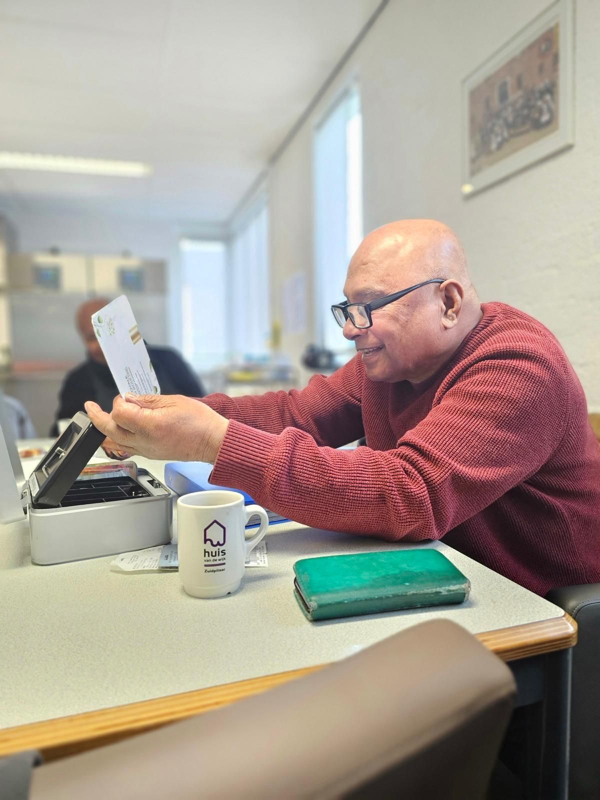 Een man in een rode trui zit aan een tafel en kijkt naar een vel papier.