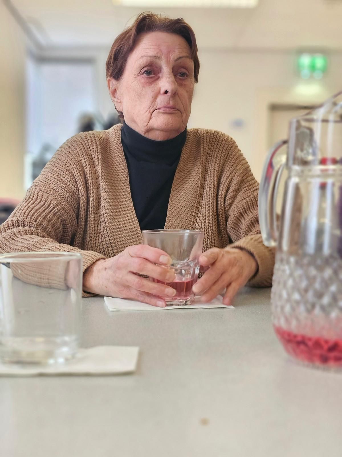Een oudere vrouw zit aan een tafel met een glas wijn in haar hand.