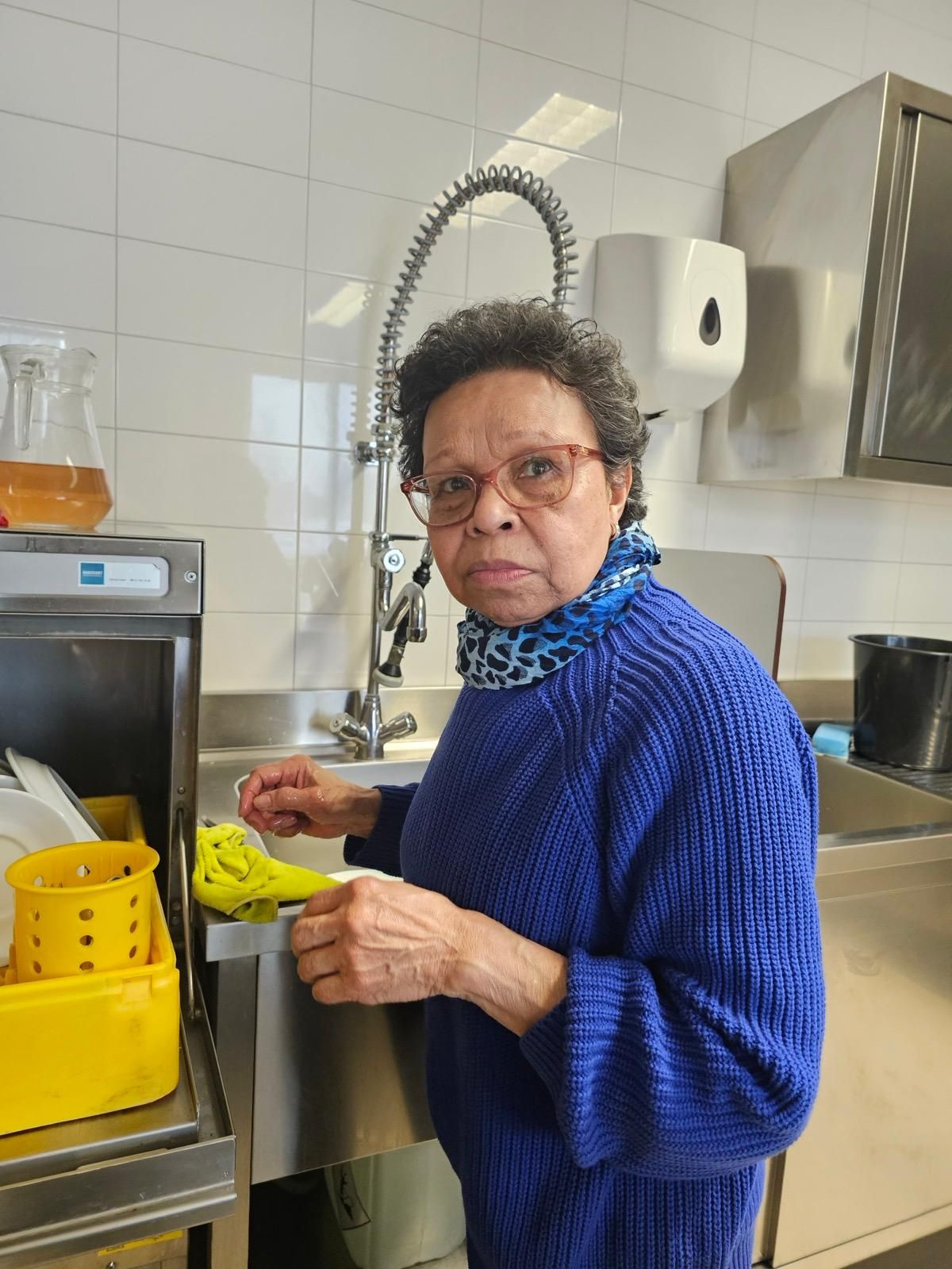 Een vrouw in een blauwe trui wast de afwas in een keuken.