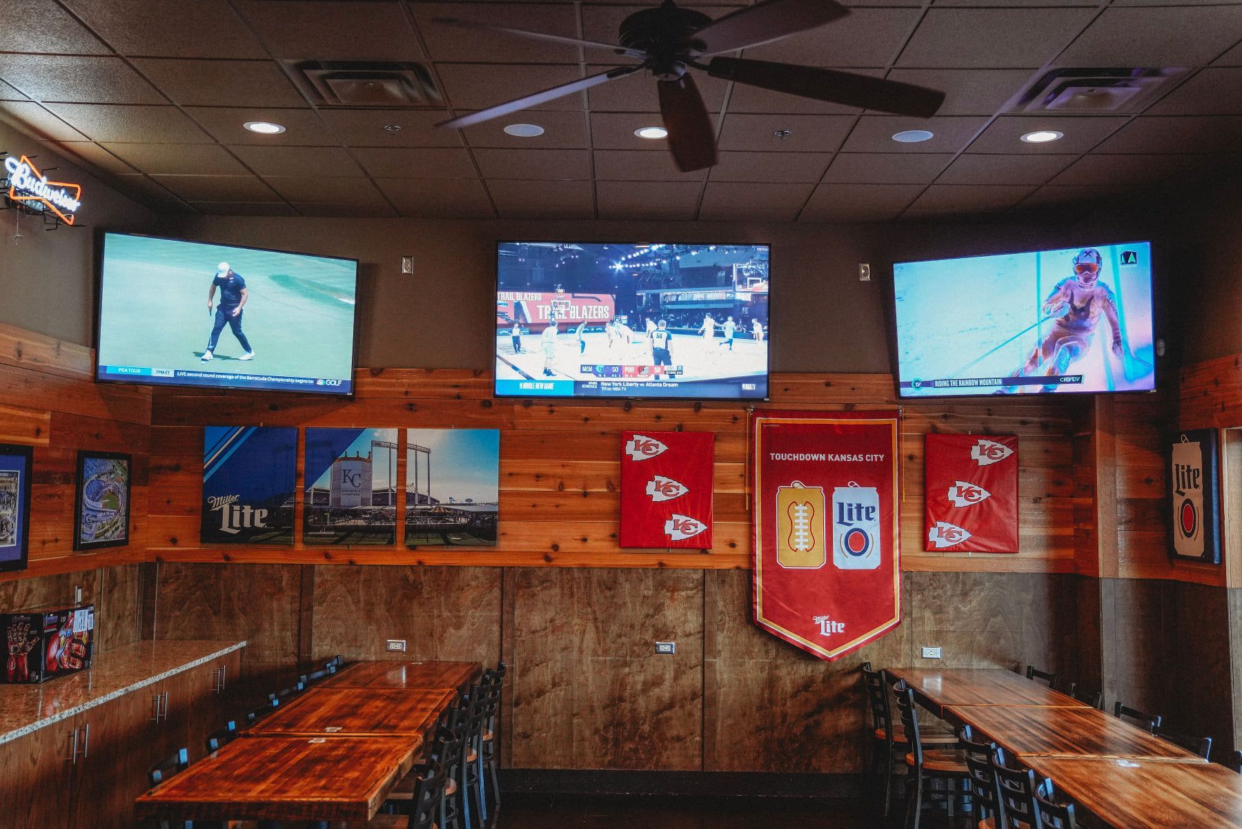 Sports bar interior with multiple TVs displaying sports. Wooden tables and a ceiling fan are visible.