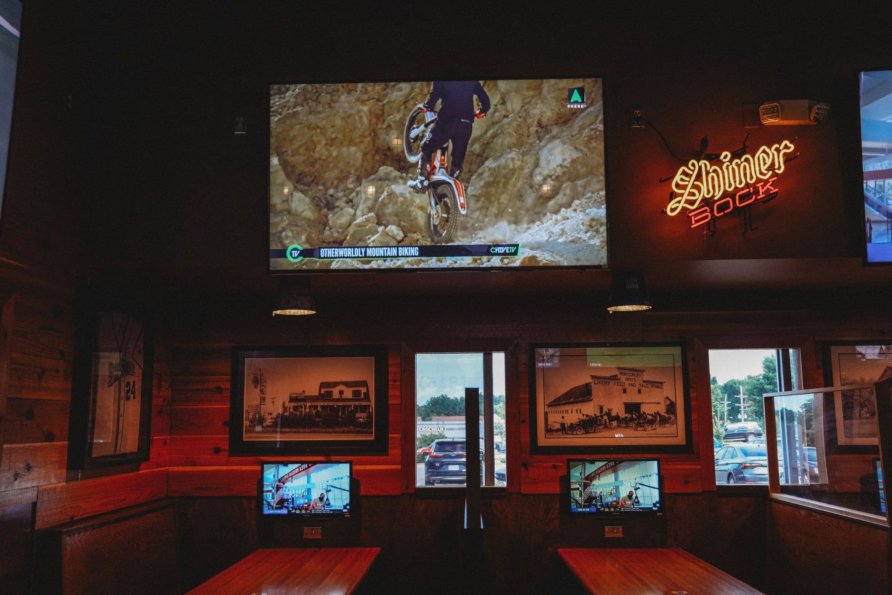 Interior of a bar, screens showing mountain biking, booths, neon sign.